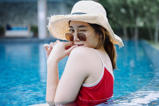 美丽的亚洲女游客戴着墨镜和帽子在游泳池里放松。