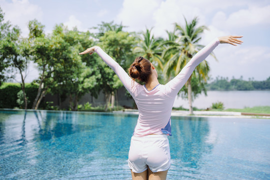 后视图-美丽的亚洲女游客伸展双臂在空中游泳池。