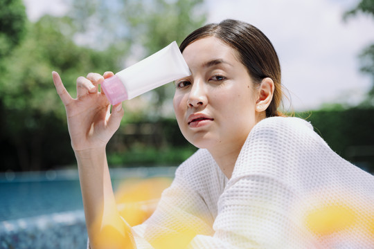 身着白色浴袍的亚洲女子在室外游泳池展示防晒乳液瓶。