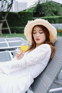 美丽的亚洲女游客戴着帽子在沙滩椅上享受着喝橙汁的乐趣。