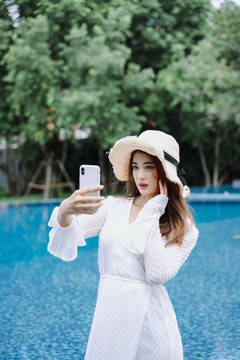 美丽的亚洲女游客，戴着帽子自拍或在游泳池用智能手机聊天。