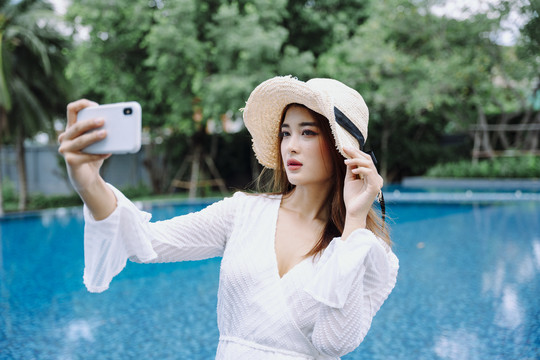 美丽的亚洲女游客，戴着帽子自拍或在游泳池用智能手机聊天。
