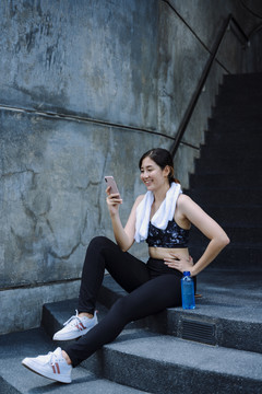 美丽的亚洲女人运动后放松，在户外楼梯上使用智能手机。