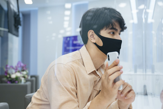 侧视图-亚洲男子戴着口罩，在美容诊所向接待员展示智能手机上的照片。