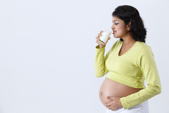 微笑着的孕妇站着，拿着一个杯子，杯子里的牛奶被隔离在灰色的背景上。