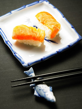 两块三文鱼寿司放在一个方形的盘子里，一双筷子放在鱼形的筷子架上