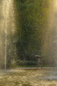 夏天水池喷泉边走过的打伞行人