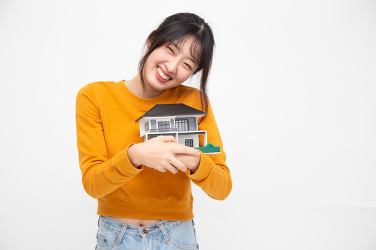 年轻的亚洲女性微笑着拥抱着梦想之家，在白色背景、房地产和家庭保险概念下孤立的样本模型
