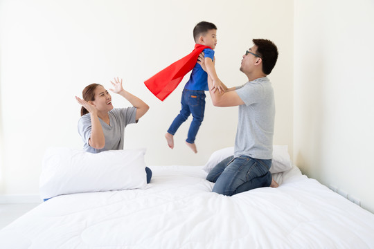亚裔父亲、母亲和儿子在卧室的床上扮演超级英雄。友好的家庭玩得开心