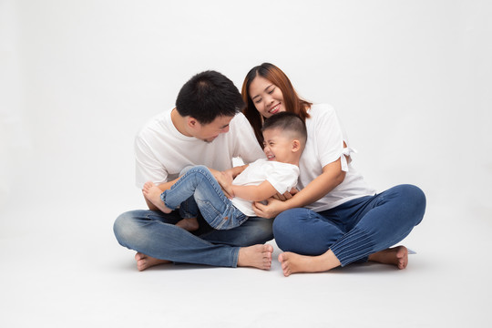一个快乐的亚洲家庭坐在白色背景下，享受父亲搔痒小儿子的乐趣。有孩子的年轻夫妇穿着白色上衣和蓝色牛仔裤