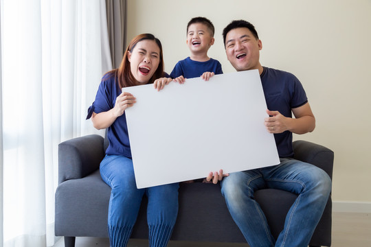 快乐微笑的亚洲家庭拿着空白的白色大海报，坐在客厅的沙发上，哇，惊喜的概念