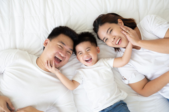 俯视图：可爱的小男孩和他的亚洲父母躺在家里的白色床上，看着相机，微笑着，幸福的家庭观念
