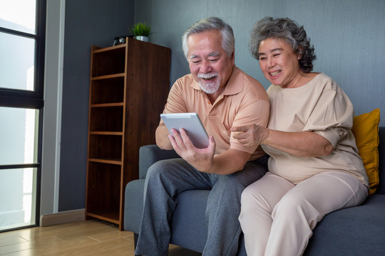 一对亚洲老年夫妇面带微笑，看着家里沙发上的同一块平板电脑，成熟的退休男女使用新的技术概念
