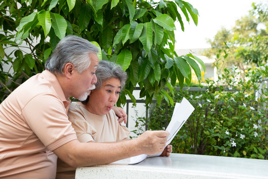 兴奋的亚洲成年夫妇在前花园家看报纸或杂志时，对惊喜的好消息感到惊讶