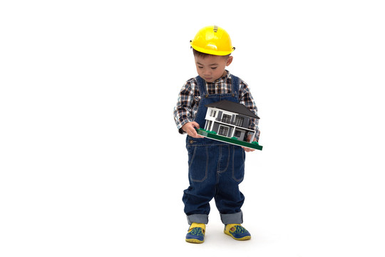 一名亚裔泰国男婴，身穿工程服，头戴安全帽，手拿白色背景隔离的样板房，房地产概念建筑工人，一岁六个月大