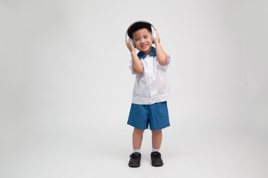 快乐微笑的亚洲小男孩，穿着学生制服，戴着白色背景上的隔离耳机听音乐，幼儿园第一天和返校概念，泰国男孩