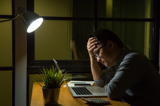 一名年轻的亚洲男子坐在桌子上，在深夜看着笔记本电脑，在办公室里感到严肃的思考和紧张。加班和努力工作的