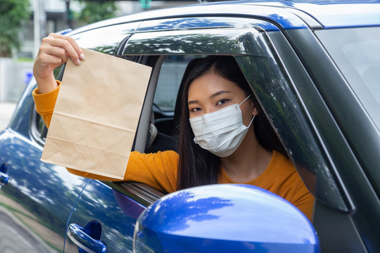 一名亚洲妇女戴着口罩，从车窗里拿着一袋快餐。免下车餐饮服务理念