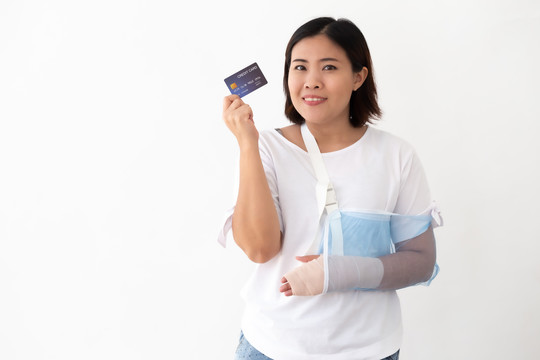 一名亚洲妇女手持信用卡，因手臂骨折而戴上软夹板，手臂被隔离在白色背景上，这是个人意外的概念
