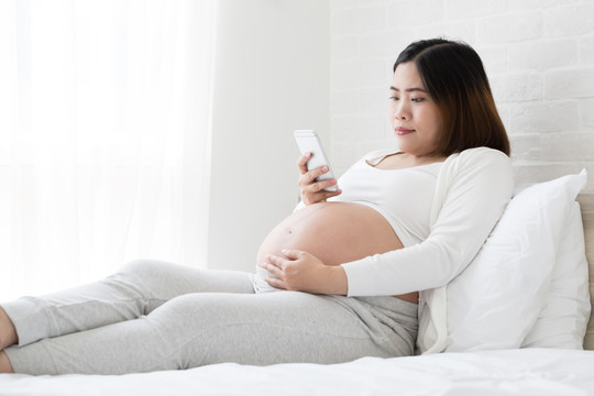 亚洲孕妇使用智能手机，在家里的床上保持最新信息，怀孕期间使用手机和社交媒体成瘾概念