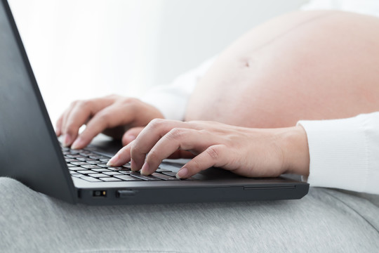 孕妇在笔记本电脑上的键盘上手工打字，搜索新生儿物品，为生儿育女做准备。怀孕期间使用笔记本电脑与技术概