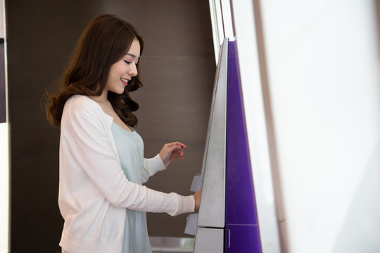 年轻的亚洲女性在自动取款机前用信用卡取款，女性站在银行的自动取款机前