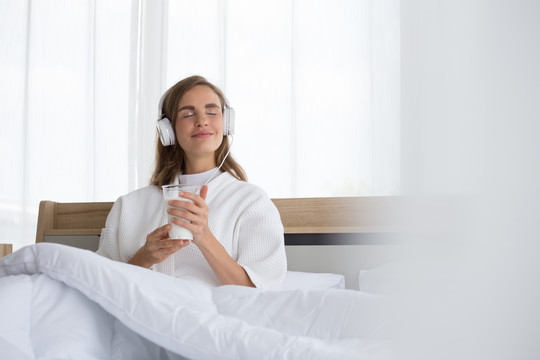早上，在公寓房间的床上，一位女士拿着一杯牛奶，听着音乐应用程序播放的放松歌曲