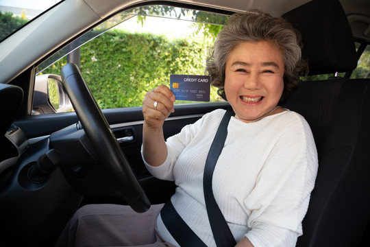 快乐的亚洲老妇人坐在车内，拿着信用卡付油费、付轮胎费、车库维修费、加油站加油费、汽车融资