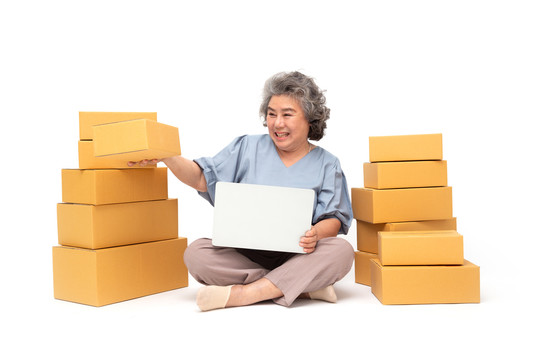 亚洲资深女性创业小企业自由职业者，手拿包裹盒和笔记本电脑，坐在白色背景的地板上，在线营销包装盒交付概