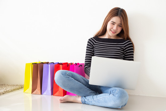 年轻的亚洲女性坐在地板上，在家里客厅里用笔记本电脑在线购物