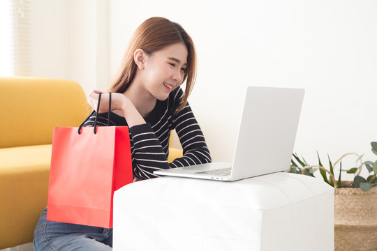 年轻的亚洲女性拿着红色纸袋，在家里客厅里用笔记本电脑上网购物