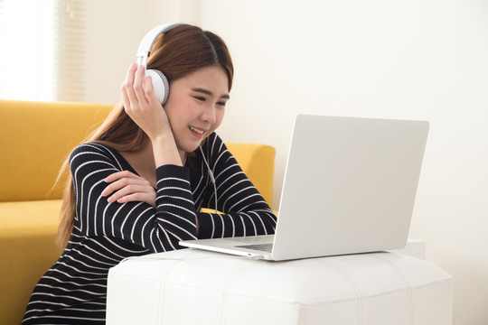 年轻的亚洲学生使用笔记本电脑，坐在客厅的地板上，用电子耳机或耳机收听和观看在线课程。互联网E-Lea