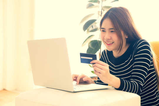 亚洲年轻女性手握信用卡，在家里的房间里用笔记本电脑进行网上购物。青少年在网上购买销售产品的概念。复古