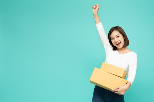 快乐的亚洲女性手持包裹盒，双手在空气中庆祝成功，背景为浅绿色，传递快递和运输服务理念