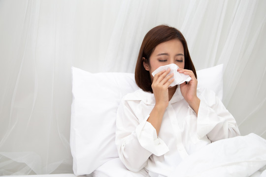 亚洲年轻女性在卧室白色床上用纸巾打喷嚏，流感和过敏概念
