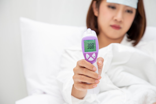 亚洲年轻女子高烧，手持数字温度计，睡在家中的白床上，病征包括发烧、咳嗽和喉咙痛，或因感染细菌或新病毒
