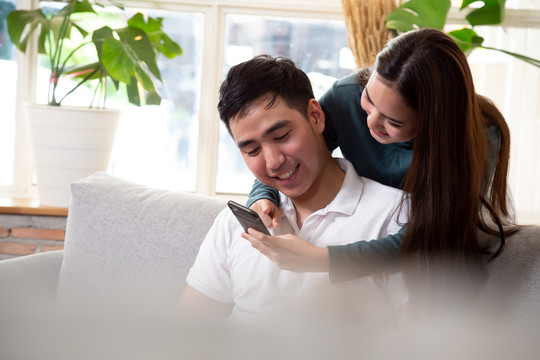 一对幸福的亚洲夫妇坐在家里的沙发上，通过手机享受媒体内容，男女朋友的关系爱情概念