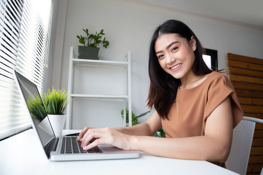 穿着休闲服的年轻亚洲女性在客厅室内工作时，使用笔记本电脑查看电子邮件，面带微笑。在家工作还是呆在家里
