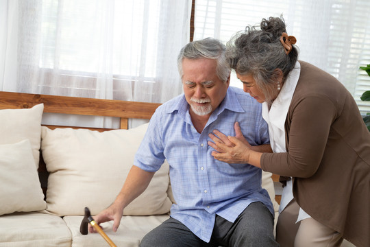 亚洲老年男性，心脏疼痛，躺在客厅沙发上，由妻子照顾，健康问题和感觉不舒服