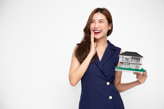 年轻的亚洲女性面带微笑，手持白色背景、房地产和家庭保险概念上孤立的房屋样本模型