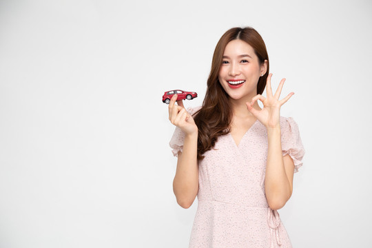 年轻的亚洲女性手持红色汽车模型，在白色背景上显示ok标志，旅游保险理念