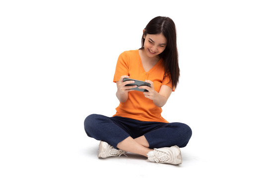 年轻的亚洲女性坐在地板上，一边用智能手机在白色背景上玩游戏，一边表现出令人惊叹的表情