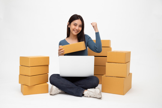 年轻的亚洲女性创业小企业自由职业者，手拿包裹盒和笔记本电脑，坐在白色背景的地板上，在线营销包装盒交付