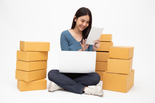 年轻的亚洲女性创业小企业自由职业者，手持平板电脑和笔记本电脑，坐在地板上，包裹盒隔离在白色背景上，在