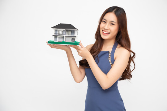 年轻的亚洲女性面带微笑，举着房子样本模型，与白色背景、房地产和家庭保险概念相隔离