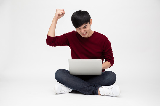 年轻的亚洲男子坐在白色背景下的地板上，拿着笔记本电脑感到很高兴