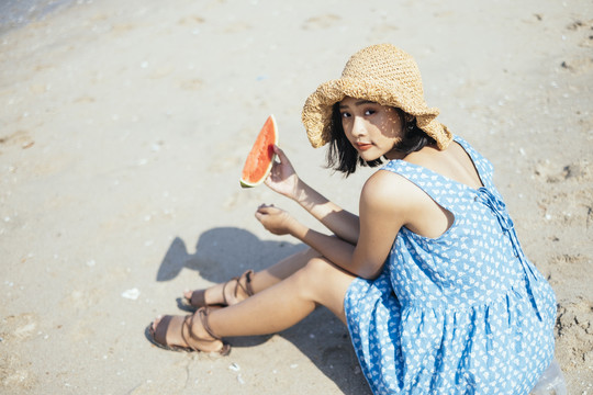 可爱的黑发女孩坐在沙滩上，手里拿着一块西瓜，对着镜头微笑。