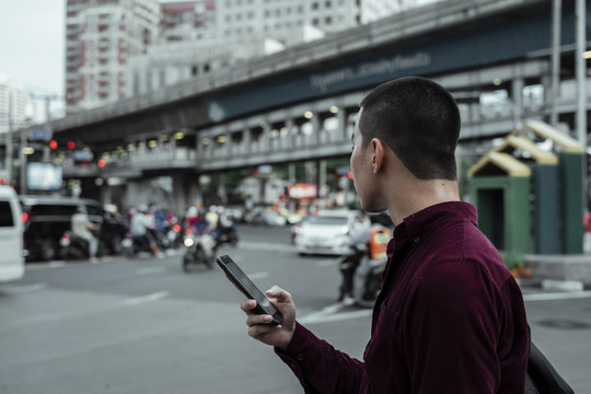 身穿红衫的光头男子在路边手持智能手机，等待穿过马路。