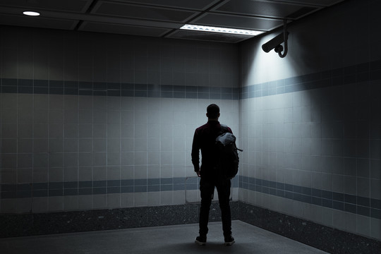 穿着红衬衫的光头男人站在黑暗的房间里，拿着一个灯光和安全摄像头，看着墙壁。