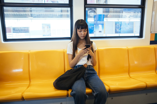 一位留着刘海发型的可爱亚洲年轻女子坐在黄色长椅上，在火车上玩手机。旅游女孩。